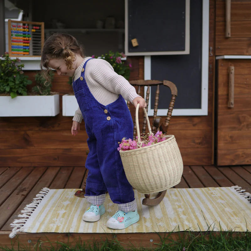 A child wearing blue overalls and a cream-colored sweater joyfully carries the Olli Ella Hand-Woven Rattan Gathering Basket in Seashell Pink, filled with flowers, while standing on a rug outside. Perfect for spring adventures and Easter activities!