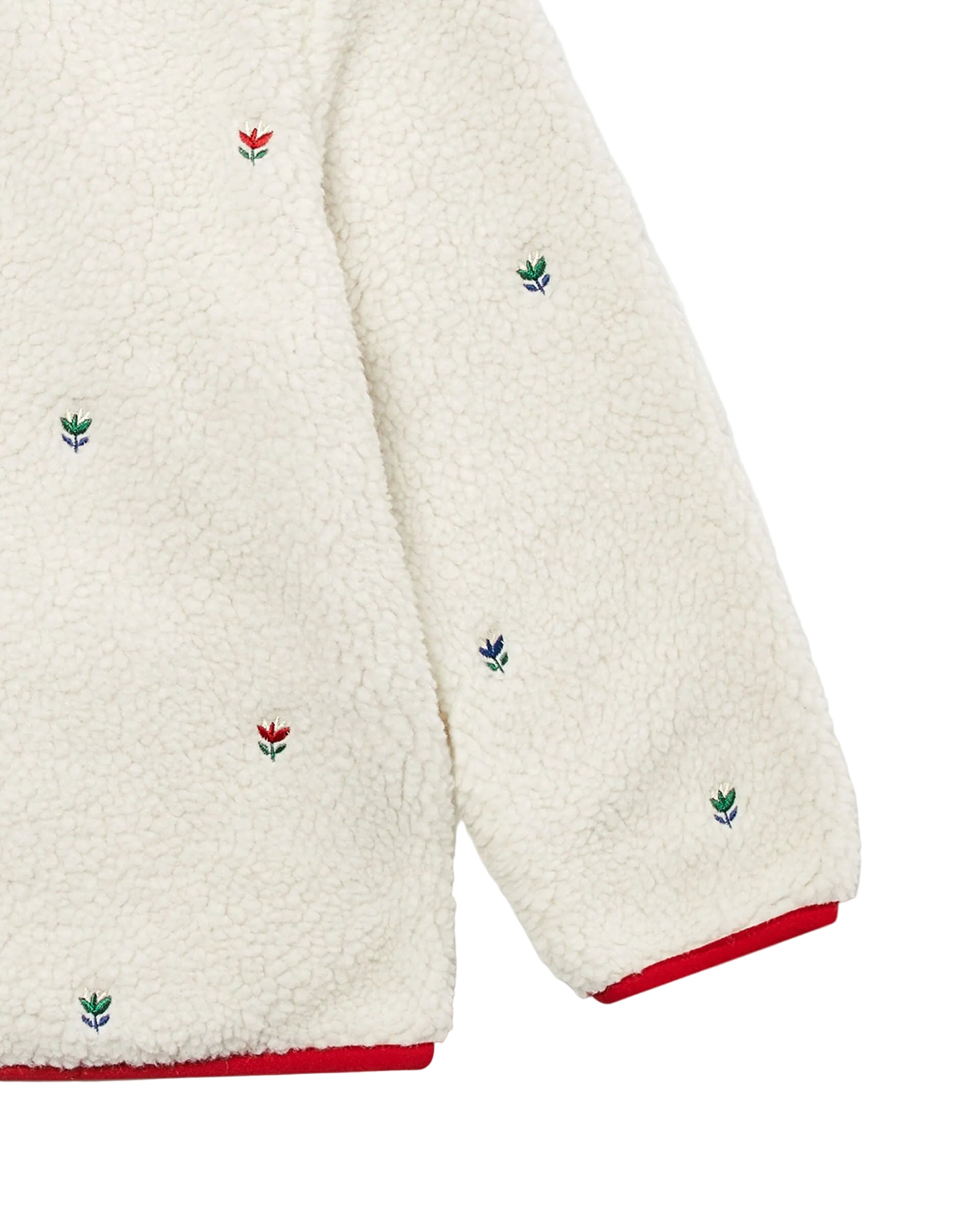 Close-up of a cream sherpa fleece garment with small embroidered floral motifs in red and green and red trim at the hem and cuff, shown on a light gray background.