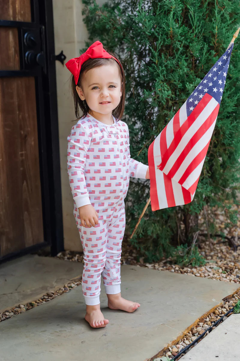 A joyful child wearing Parker Zipper Pajama American Flag by Lila and Hayes, holding a small American flag, highlighting the fun, festive spirit of these adorable american flag pajamas.