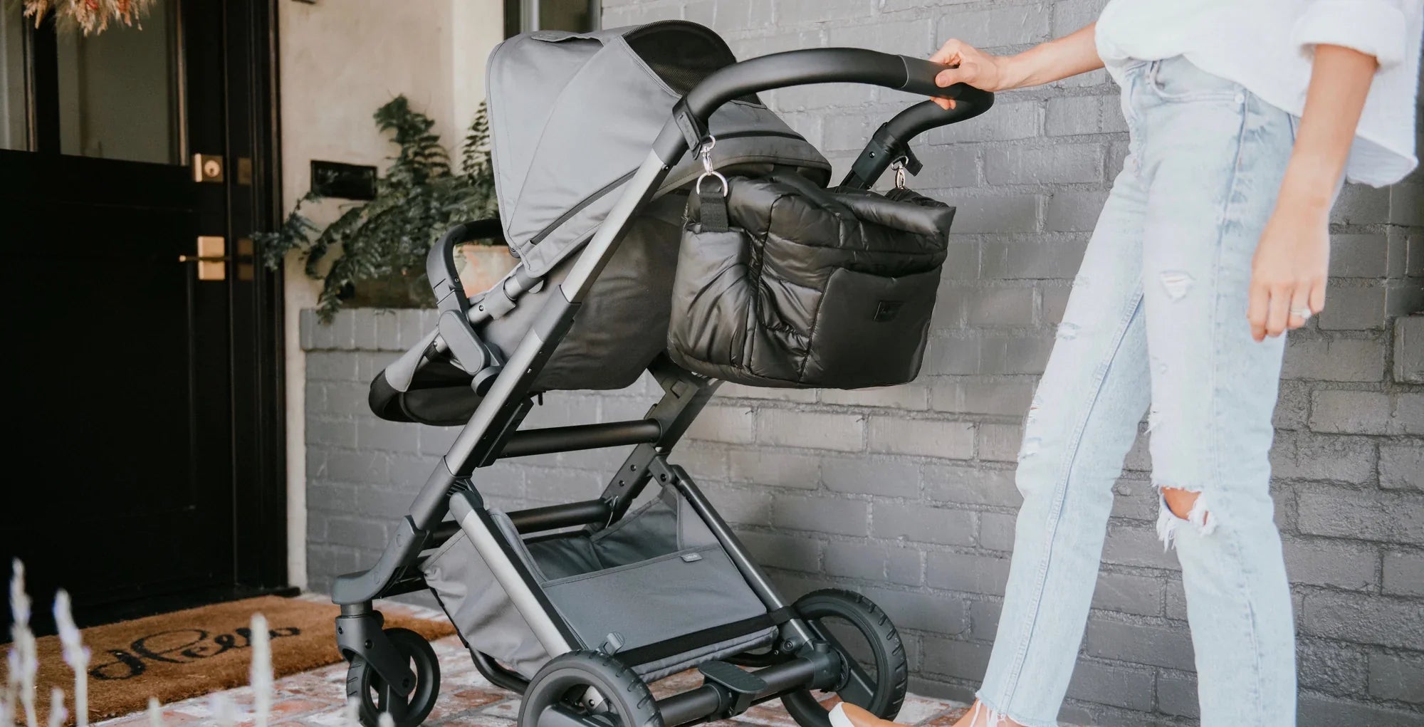 A stylish parent pushing a modern stroller featuring a cozy, quilted black accessory bag. The image showcases the 7AM Enfant brand's urban and functional design in a chic outdoor setting.