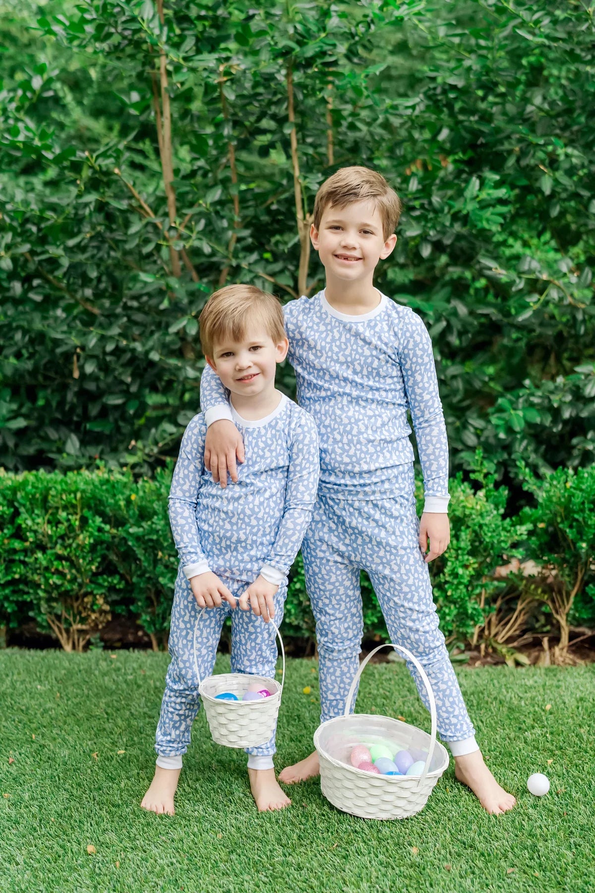 Two children wearing the Grayson Pajama Set - Easter Time Blue by Lila and Hayes, enjoying an outdoor setting with baskets, representing a playful vibe in kids cotton pajamas.