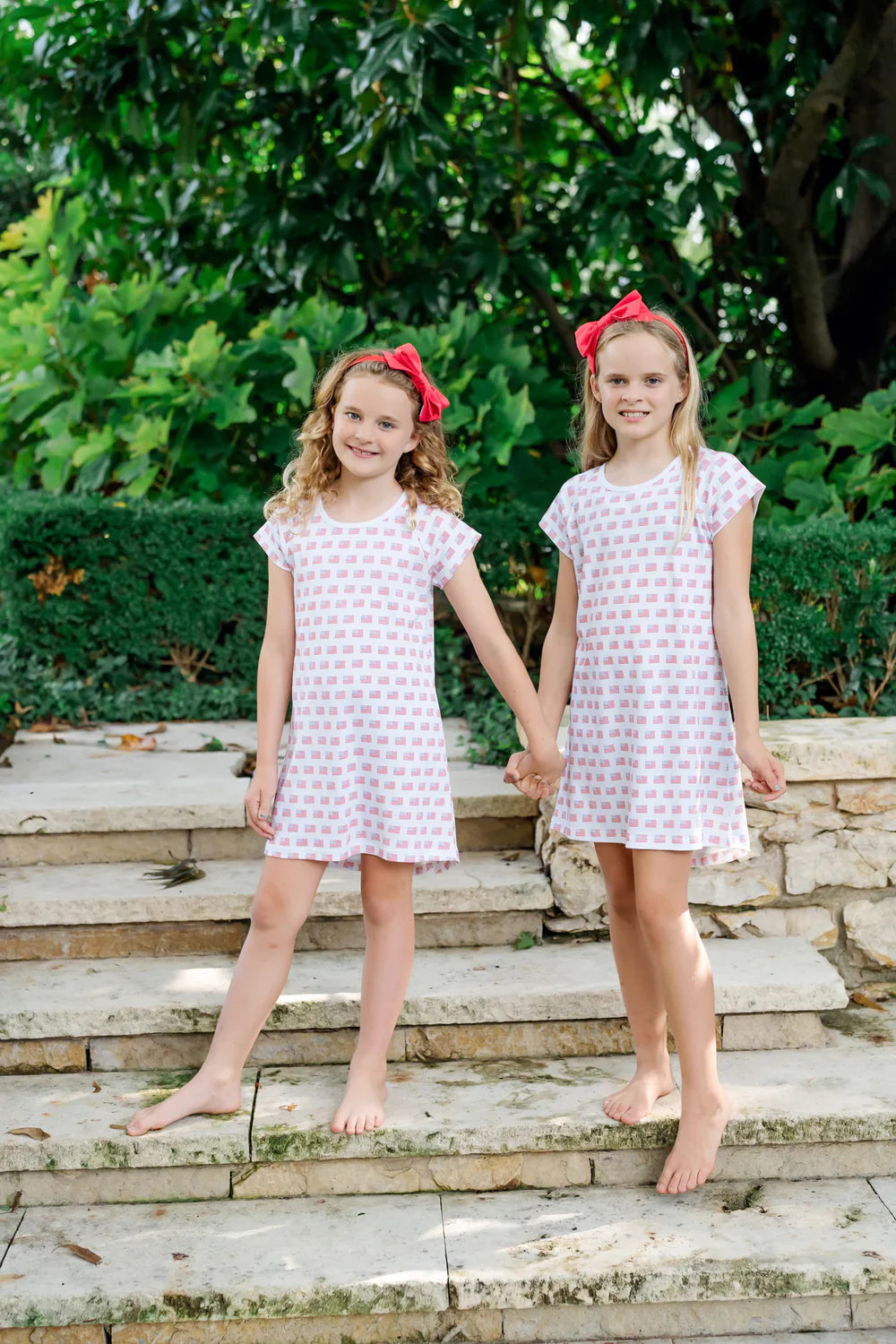 Two girls wearing the Sadie-American Flag dress by Lila and Hayes, smiling while holding hands in a lush garden setting.