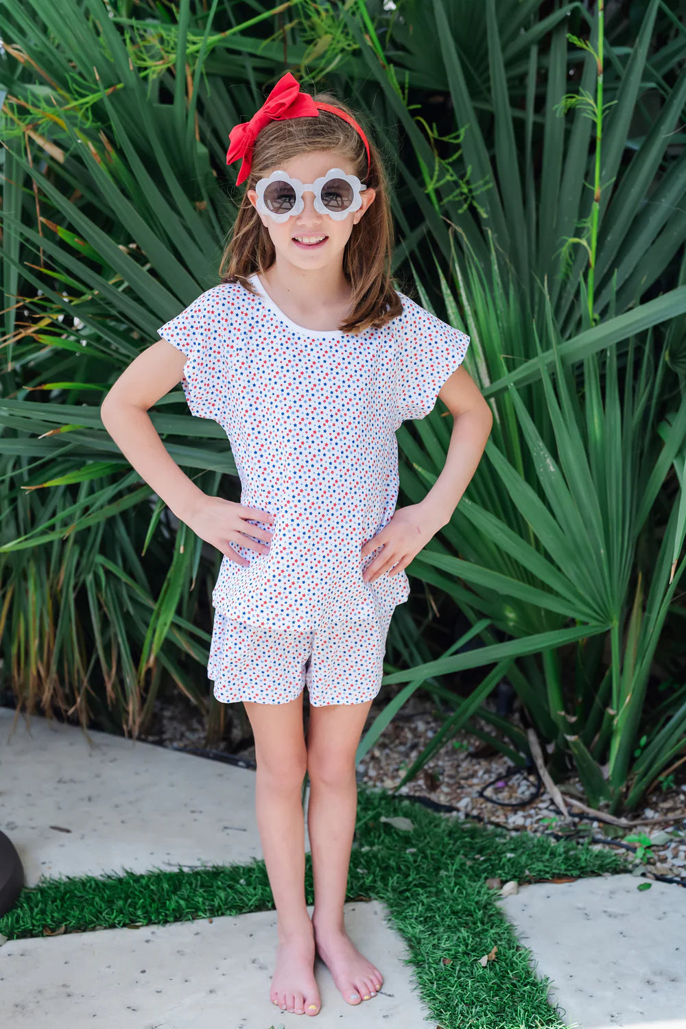 Child wearing the Emery-Star Spangled short cap sleeve top and short set by Lila and Hayes, posing outdoors with playful sunglasses and a bright smile.