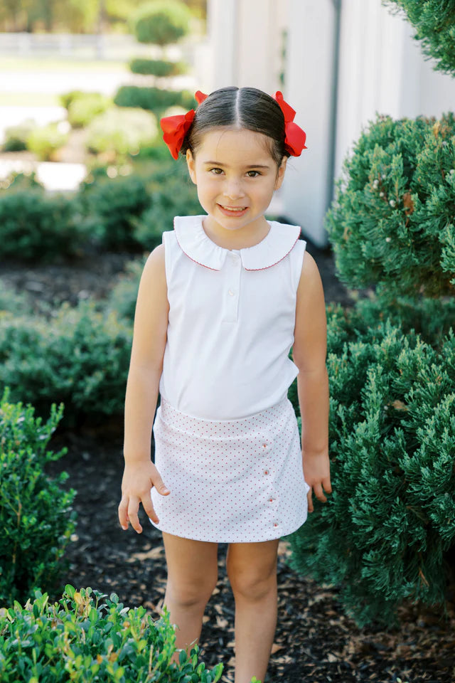 A smiling child wearing the Dottie Skort from The Proper Peony, paired with a white top and red hair ribbons, set against a backdrop of green bushes, perfect for a cheerful summer look.