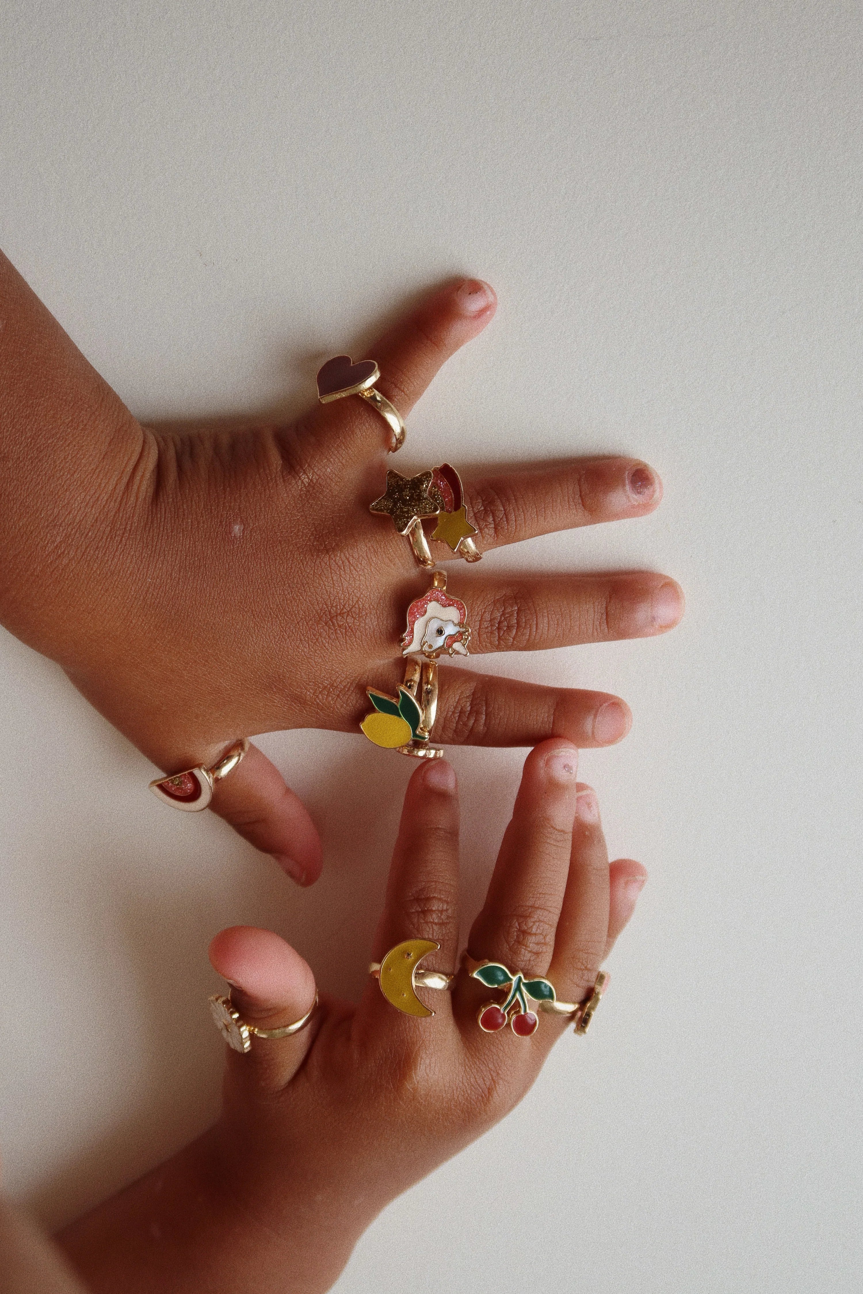 Two hands filled with various rings from the 12 Pack Ring Set from Konges Slojd, each ring showcasing different whimsical motifs.